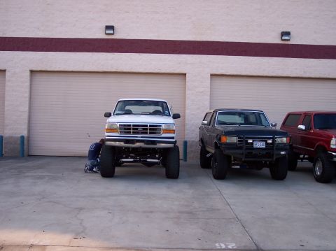 1995 Ford Bronco - Three lifted Ford trucks, including a white F-150 and two Broncos, are parked in front of garage doors.