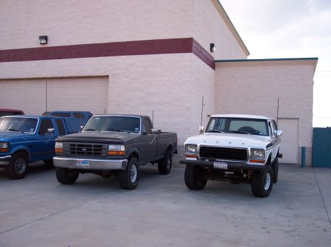 1995 Ford Bronco - A white 1978 Ford Bronco is parked next to two trucks, one dark gray and one blue, outside a commercial building.