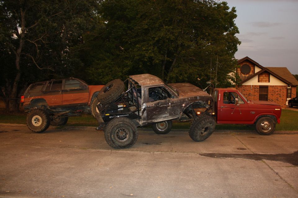 Modified brown pickup truck with roll cage and spare tire, parked on a street next to other vehicles