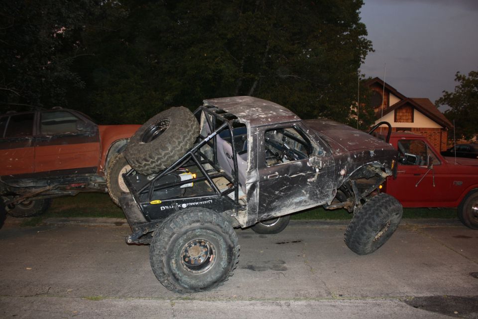 Modified black truck with roll cage and large tires, parked next to a red truck and an orange vehicle