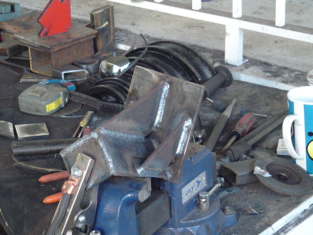 Welded steel bracket clamped in a blue bench vise on a cluttered workbench