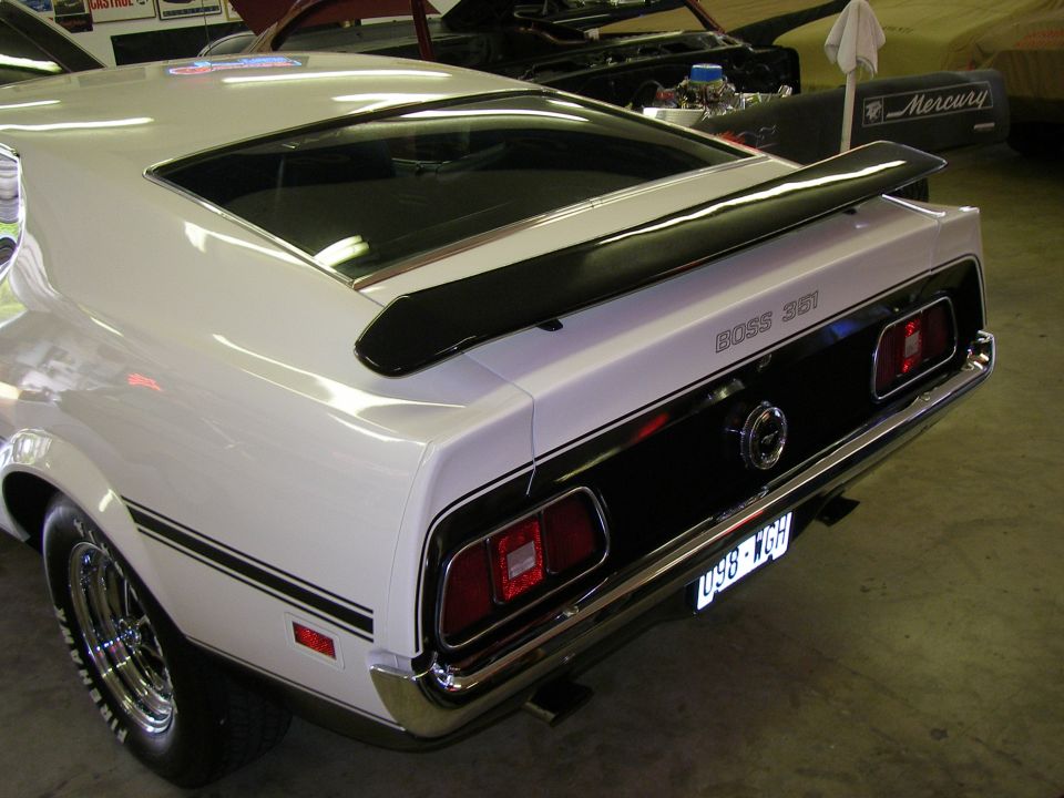 Rear view of a white 1971 Ford Mustang Boss 351 with black stripes and spoiler. Another car is visible in the background