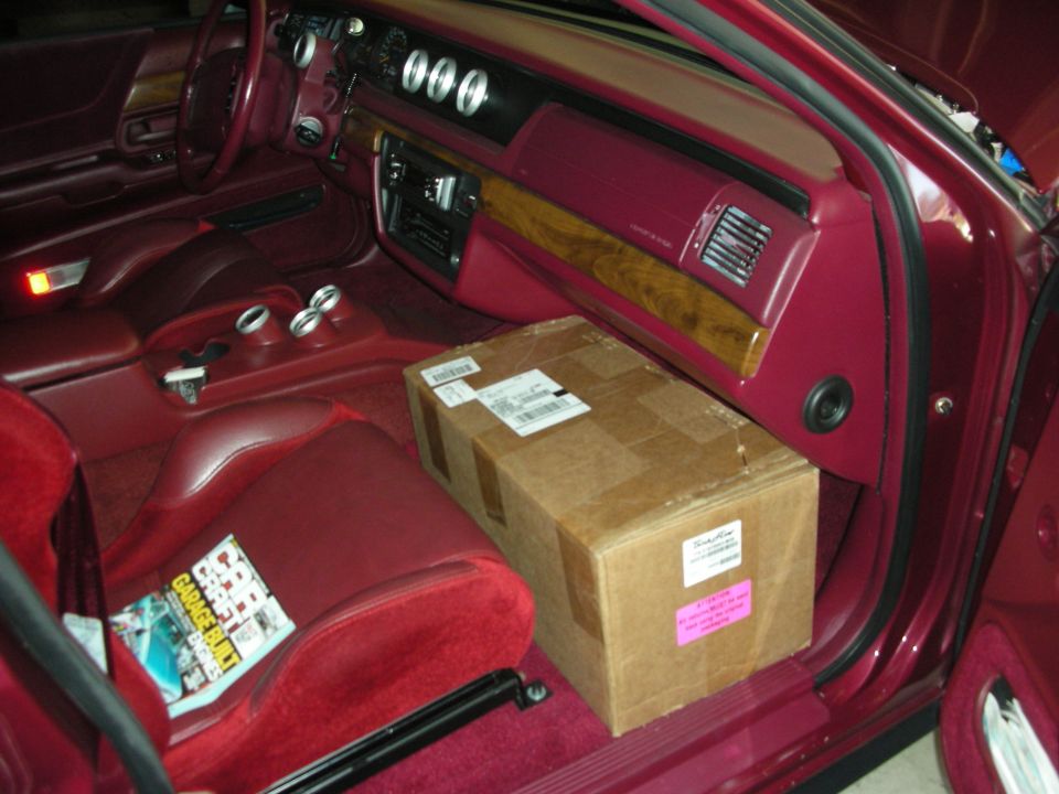 Car interior with maroon seats and carpet. A cardboard box sits on the passenger seat. A magazine is on the driver's seat