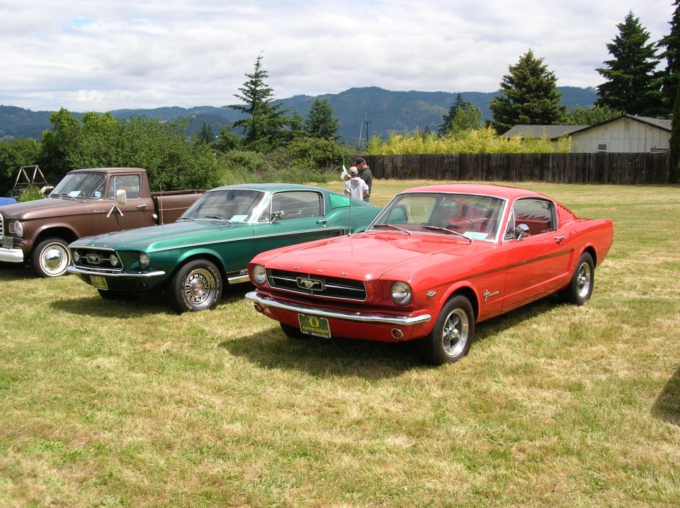 Red and green classic Ford Mustang fastbacks parked on grass at a car show, hills in background
