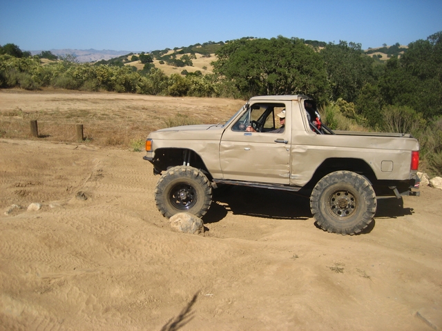 Tan topless Ford Bronco, side profile on a dirt trail, lifted on mud-terrain tires, rolling hills