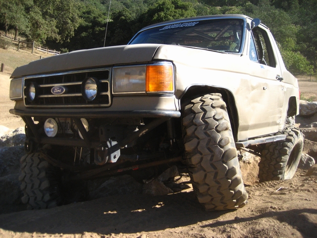 Tan chopped-top Ford Bronco on tall mud tires climbing a rock ledge, low front angle