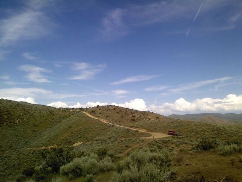 1981 Ford Bronco - A red SUV navigates a dirt road winding through rolling, brush-covered hills under a vast sky.