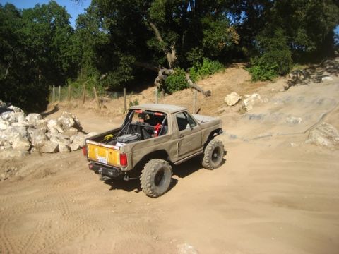 1989 Ford Bronco - This modified SUV is navigating a dusty, off-road trail with large boulders and trees nearby.