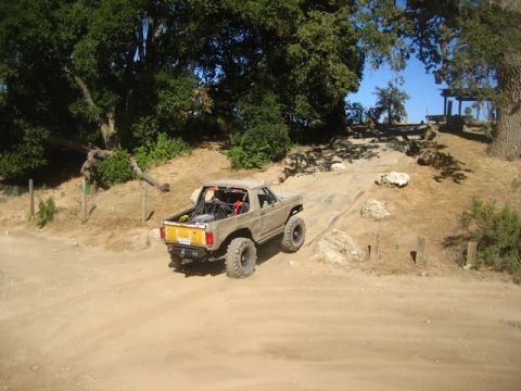 1989 Ford Bronco - A tan Ford Bronco navigates an off-road trail with large tires and a roll cage visible.