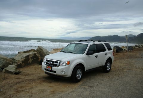 1996 Ford Bronco - A white Ford Escape SUV is parked by the ocean under a cloudy sky, with mountains visible in the distance.