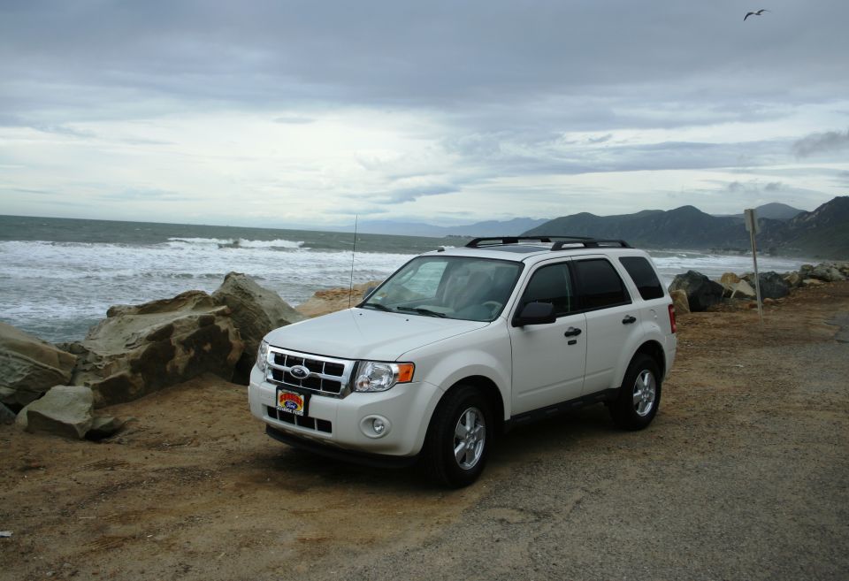 White Ford Escape SUV parked on a dirt road near the ocean, with a rocky shoreline and mountains in the background