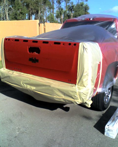 1993 Ford C-350 (Centurion Conversions) - A red pickup truck's tailgate is masked and covered for painting, with the Chevrolet emblem clearly visible.