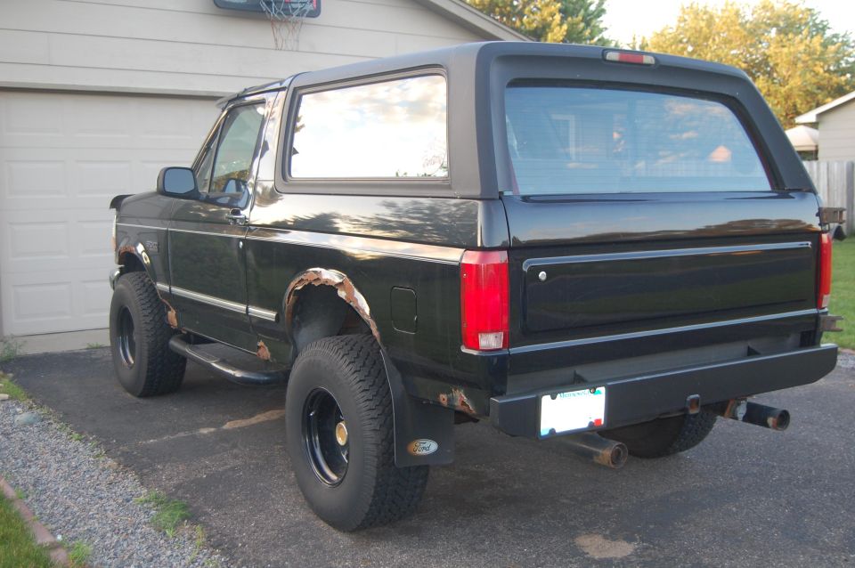 Black Ford Bronco parked in driveway, rear view. Shows rust on wheel wells and aftermarket bumper