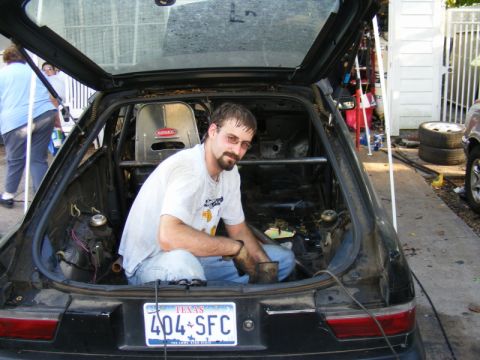 1989 Ford Escort - A man sits inside the cargo area of a modified car, which appears to be undergoing some form of customization or repair.