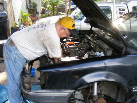 1989 Ford Escort - A man works on the engine of a black 1989 Ford Escort, with another vehicle visible in the background.