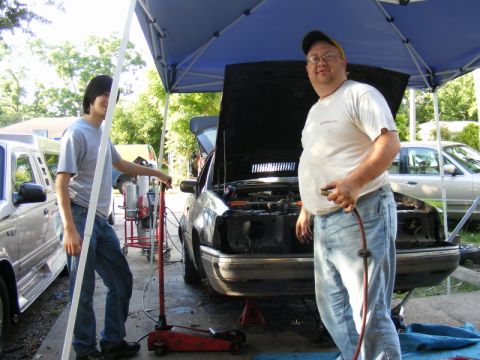 1989 Ford Escort - Two men work on a dark sedan with its hood open, parked under a blue canopy.