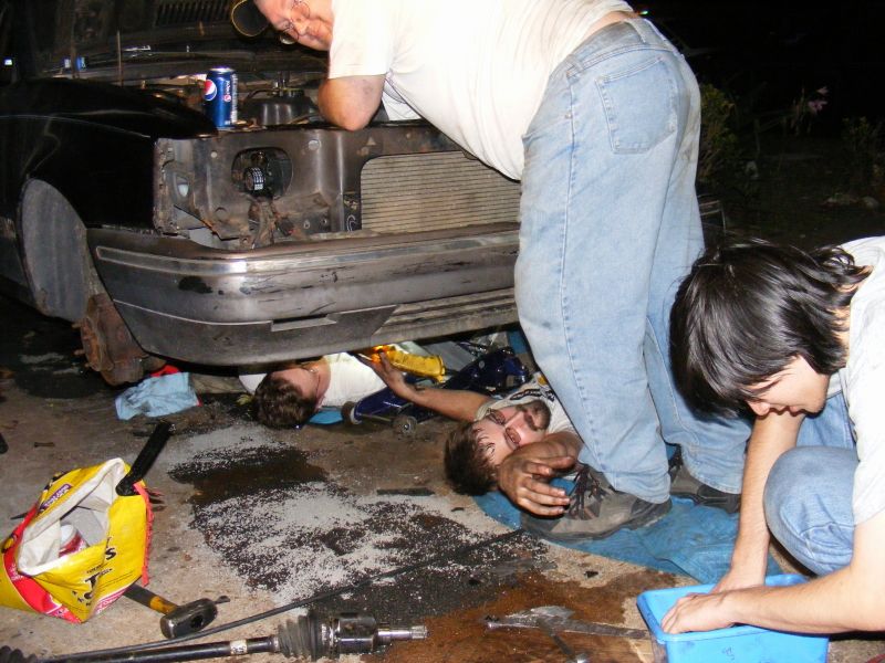Three men working on the engine bay of a car, one man is looking into the engine while two others are underneath