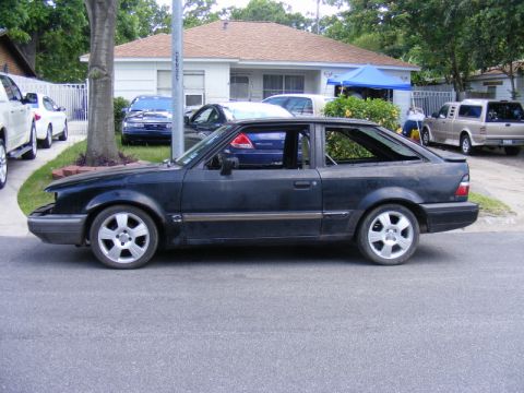 1989 Ford Escort - A black hatchback is parked on the street in front of a house with other vehicles visible.