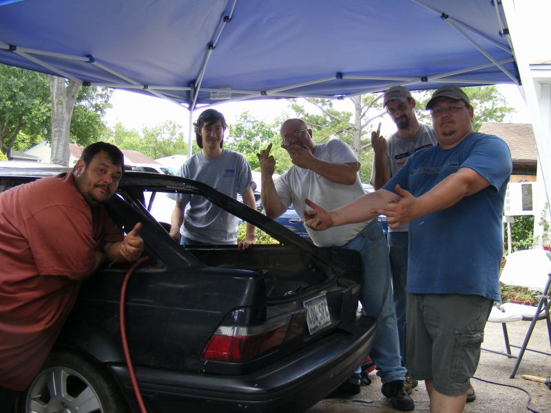 Five men gathered around the open trunk of a dark-colored 1989 Ford Escort hatchback