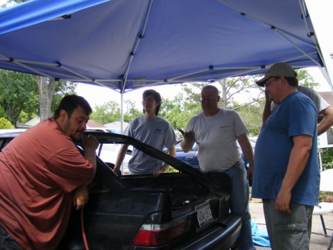 1989 Ford Escort - Several men stand under a blue canopy, looking into the open trunk of a car.