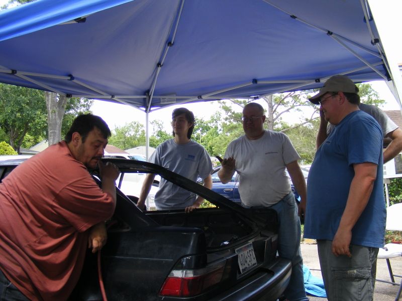 Guys gathered around the open trunk of a dark car, possibly working on it
