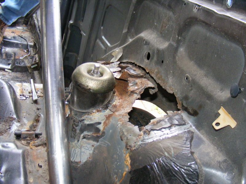 Car interior showing severe rust damage around the strut tower and floor pan. A roll cage bar is visible