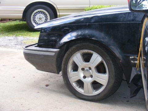 1989 Ford Escort - A black car's front fender and wheel are visible, with another car in the background.