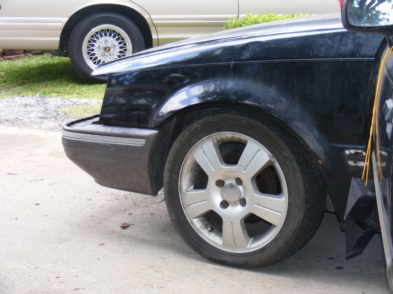 Black car front fender and wheel, with a tan car in the background. The black car has a dented fender and aftermarket wheels