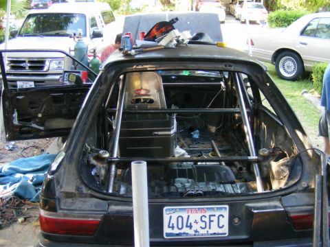 1989 Ford Escort - A modified hatchback is being worked on, with a roll cage installed and interior stripped. A Ford Ranger pickup is parked next to it.