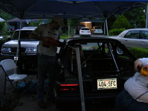 1989 Ford Escort - Two people work on a black car with a roll cage under a canopy, with other vehicles visible in the background.