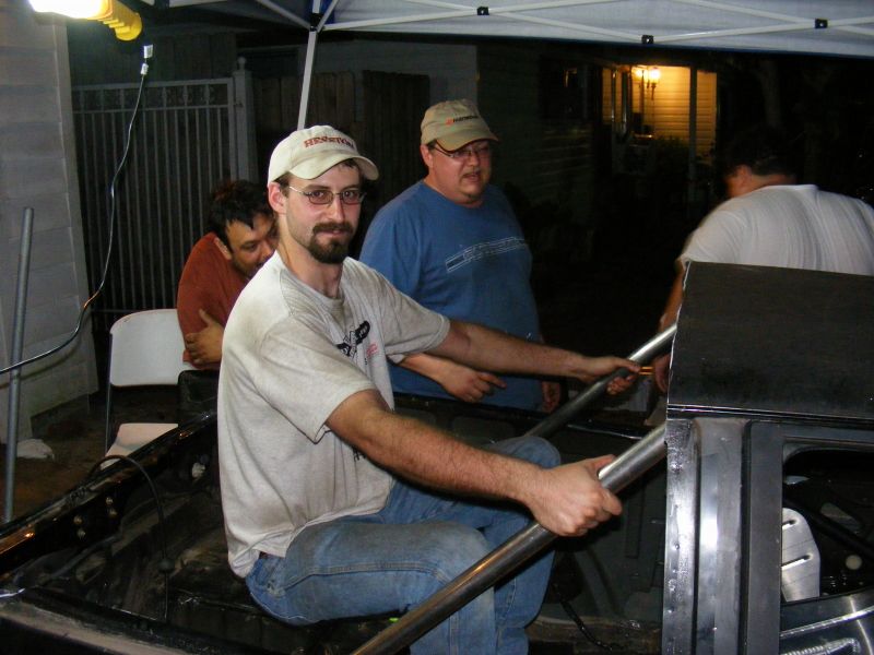 Interior of a black car with roll cage, men working on it. One man in foreground wears glasses and a cap