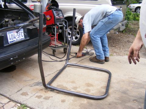1989 Ford Escort - A person works on a metal frame near the open trunk of a black car.