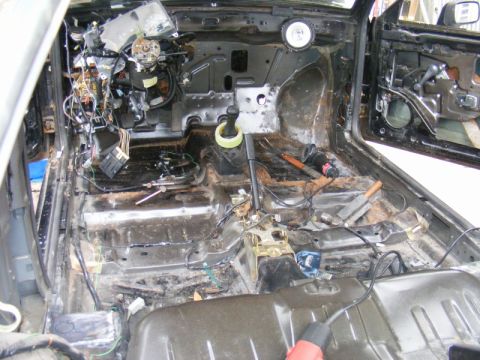 1989 Ford Escort - The interior of a car has been stripped down, revealing exposed wiring, rust, and the bare metal of the chassis.