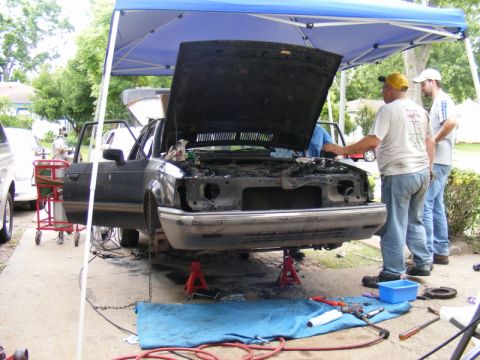 1989 Ford Escort - Mechanics work on a car with its hood up, parked under a blue canopy.