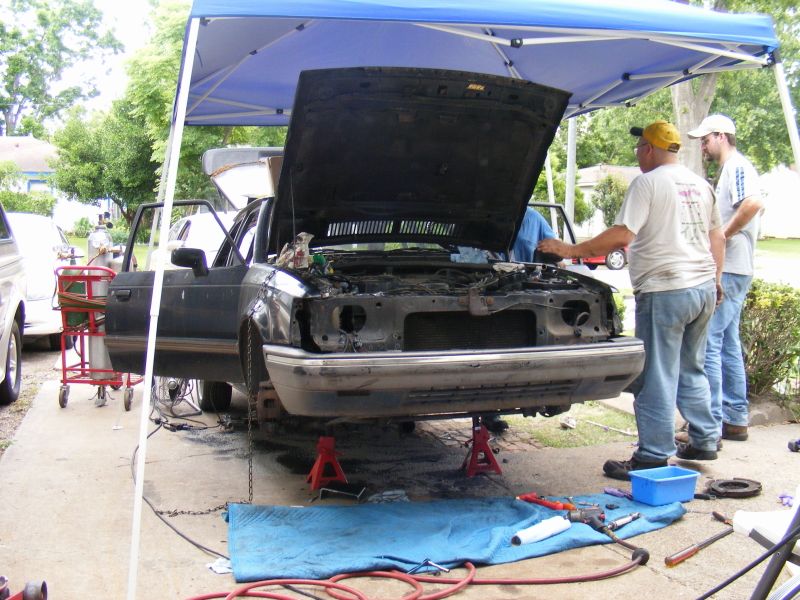 Car on jack stands with hood open, people looking at engine. Tools and blue tarp on ground