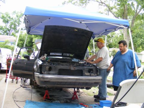 1989 Ford Escort - Two men work on a car's engine under a blue canopy, with the vehicle elevated on jack stands.