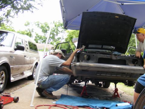 1989 Ford Escort - Mechanics work on the front end of a car under a blue canopy, with a van parked to the left.