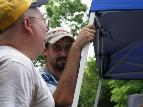 1989 Ford Escort - Two men are seen under a blue canopy, with one man in the foreground wearing a yellow cap and glasses.