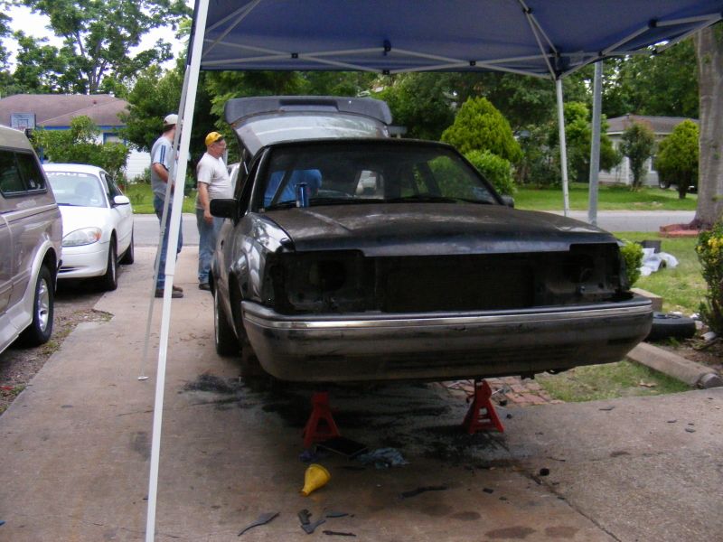 Black sedan on jack stands with hood open, two people nearby. Another white car and part of a van are visible