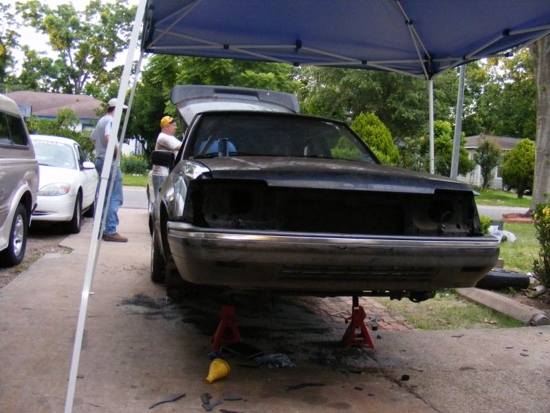 Front of a dark-colored car on jack stands, hood open, missing grille and headlights. Another white car is parked nearby