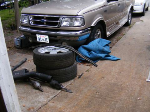 1989 Ford Escort - A tan Ford pickup truck is parked with tires and car parts on the ground in front of it.