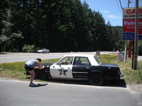 1991 Ford Bronco - A woman photographs a police car with a star emblem on its side, parked near a gas station.