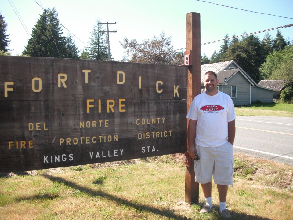 Man in white t-shirt and shorts stands by a large wooden sign that reads "FORT DICK FIRE DEL NORTE COUNTY FIRE PROTECTION DISTRICT KINGS VALLEY STA."