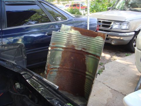 1989 Ford Escort - A rusty barrel obstructs the view of a blue car and a beige pickup truck.