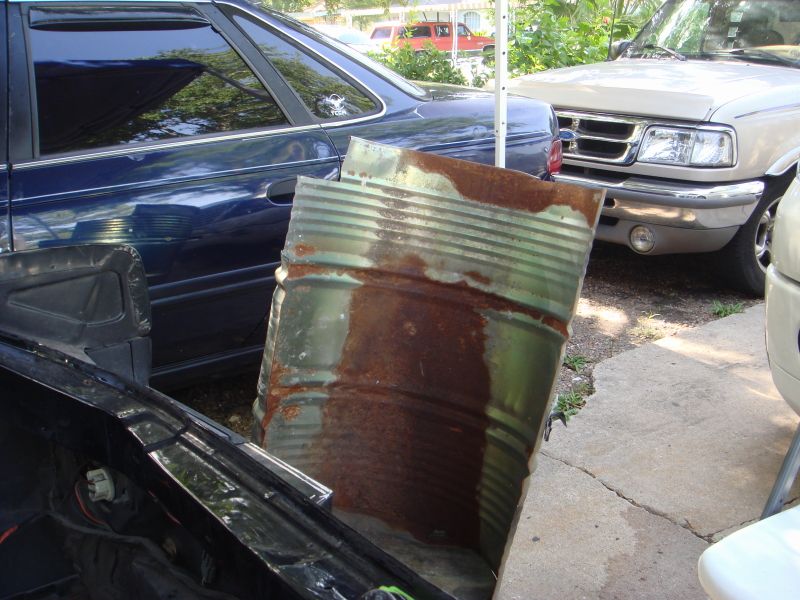 Rusty metal barrel in front of a blue car and a beige pickup truck