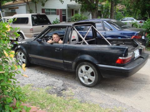 1989 Ford Escort - A child gives a thumbs up from the driver's seat of a modified black 1989 Ford Escort convertible with a roll cage.