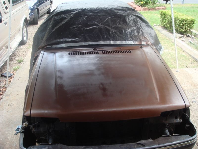 Brown car hood and front end covered by a black tarp, with another vehicle visible to the left