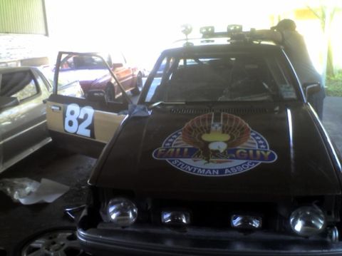 1989 Ford Escort - Customized car with 'Fall Guy Stuntman Assoc' decal and lights on roof, parked next to another car.