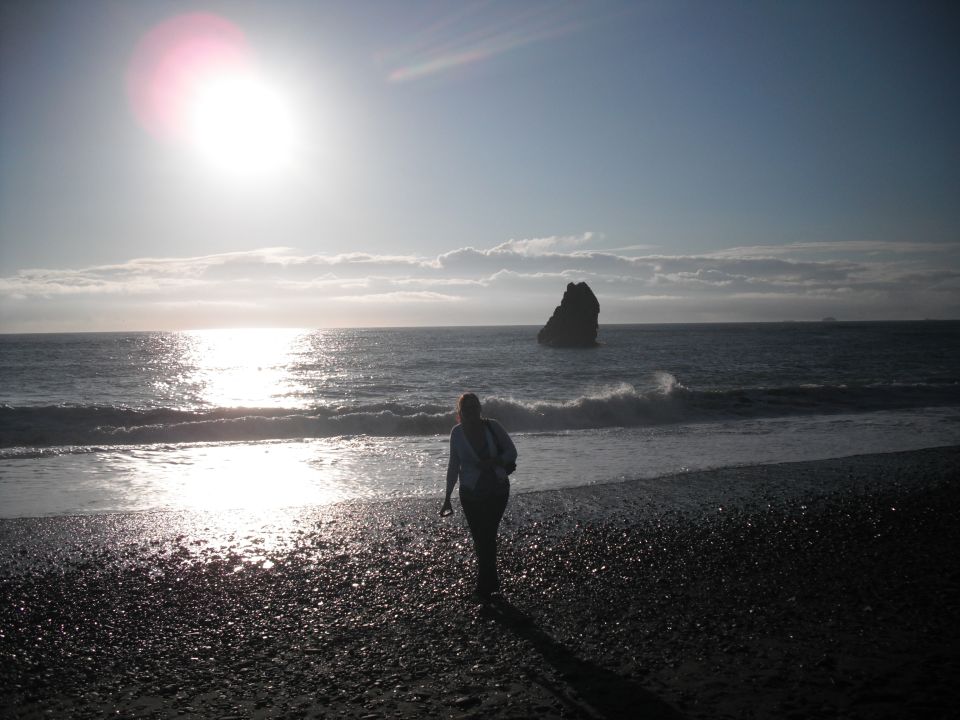 Person walks on a rocky beach toward the ocean with a large rock formation offshore