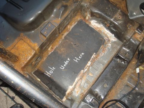 1989 Ford Escort - The interior of a heavily rusted car floor shows a handwritten note indicating a hole underneath.